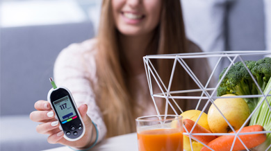 A smiling woman holding blood sugar reading device near a basket of fruits and vegetables