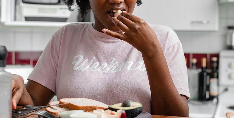 woman in light pink shirt sitting at a table with a sandwich and taking a bite